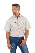 A man with short gray hair and a beard wears a beige Ringers Western Mens Lake Argyle S/S Full Button Work Shirt (118110004) and blue jeans. He stands, looking left, with one hand in his pocket.