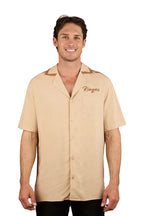 A man smiles wearing the Ringers Western Mens Carter S/S Pub Shirt (125002RW) in Dark Sand, featuring a brown collar, short sleeves, and "Ringers" embroidered on the left chest pocket. The background is plain white.