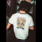 A man viewed from behind wearing a ringer t-shirt with a large vintage-style Western graphic on the back. The shirt is an off white with dark navy blue trim at the collar and sleeve cuffs, featuring a graphic of cowboys with an orange sun and the text 'Quality Western Goods Ringers Western'.