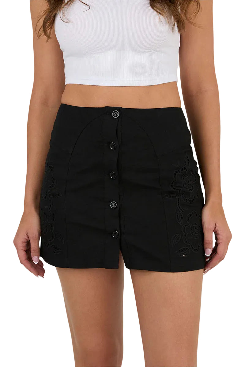 A woman wears a white sleeveless crop top paired with the Ringers Western Womens Jynette Button Down Mini Skirt in black, featuring a high-waisted fit, front buttons, and subtle floral embroidery from Ringers Western.