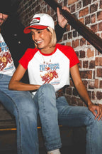 A woman wearing a red and white raglan t-shirt with a 'Bull Riding Champion' graphic, paired with a matching baseball cap, sitting on a staircase next to a brick wall.