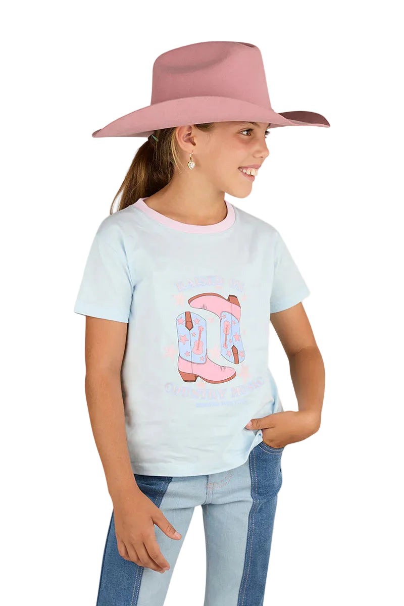 A young girl in a pale pink cowboy hat, blue jeans, and the Ringers Western Girls Raised On Country Music Classic Fit S/S Tee (Barely Blue) smiles and looks off to the side with one hand in her pocket against a white background.