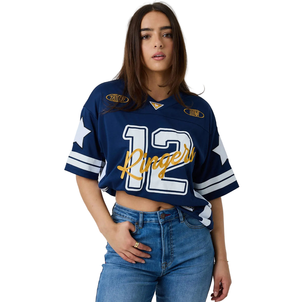 A woman modeling a navy blue sports jersey tucked up with '12 Ringers' in white and gold script, paired with blue jeans, against a white studio background.