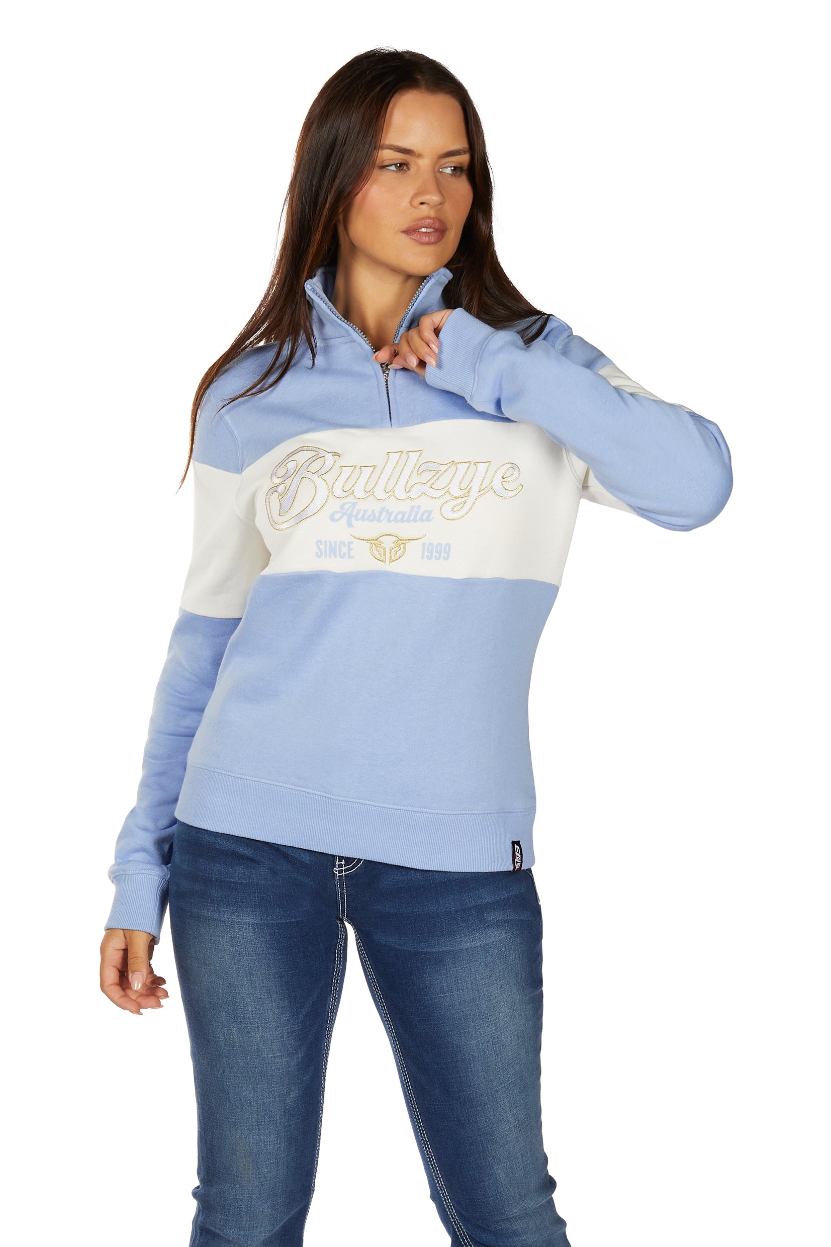 A woman in blue jeans models the Bullzye Womens Kai 1/4 Zip Pullover (B6W2597531) in light blue/cream, featuring "Since 1999" text. This stylish Bullzye pullover offers a cozy blend of comfort and fashion.