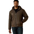 A man stands facing forward, one hand in his pocket, wearing the Ariat Mens F20 Rebar Duracanvas Jacket (10027842) in Wren. The brown, hooded canvas outerwear with visible stitching pairs well with his black shirt and blue jeans.