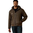 A man stands facing forward, one hand in his pocket, wearing the Ariat Mens F20 Rebar Duracanvas Jacket (10027842) in Wren. The brown, hooded canvas outerwear with visible stitching pairs well with his black shirt and blue jeans.