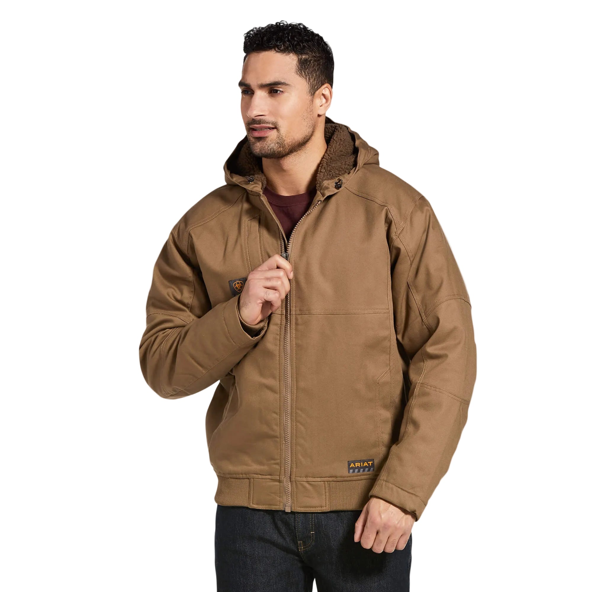 A man with short curly hair is wearing an Ariat Men's Rebar Duracanvas Jacket (10032964) in Field Khaki and dark jeans. He is facing slightly left, looking forward, and zipping up the front of the Ariat jacket.