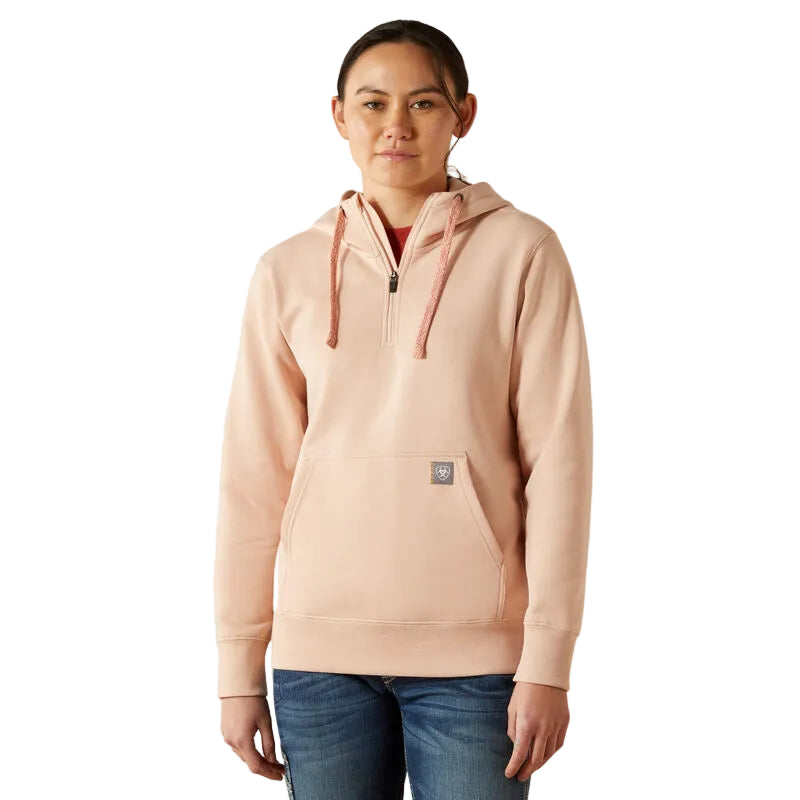 A woman faces forward in an Ariat Womens Rebar Skill Set 1/2 Zip Hoodie (10062652) in Cameo Rose—a light pink, water-repellent hoodie with a front pocket—against a plain white background.