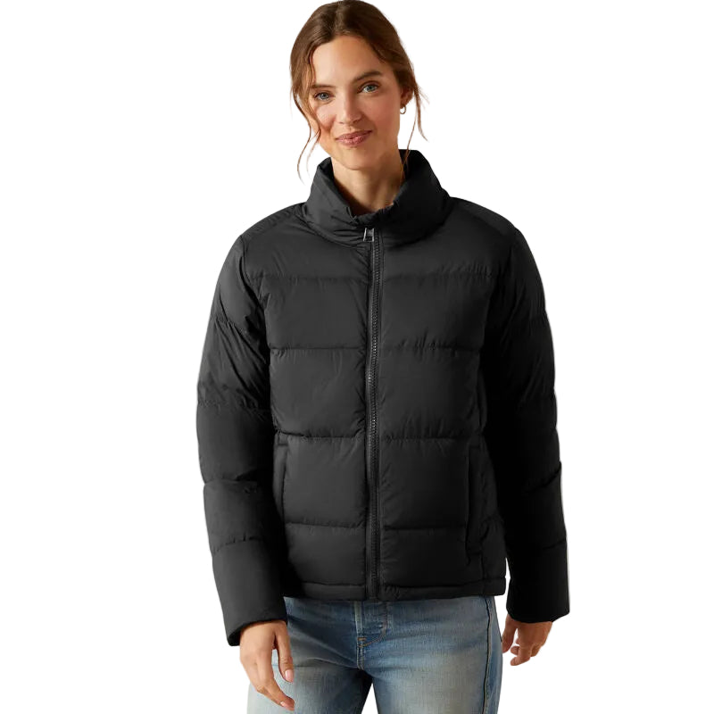 A person stands smiling in light blue jeans and the Ariat Womens Ultrapuff Down Jacket (10065822) in black by Ariat, featuring a water-repellent finish and front zipper, set against a plain white background.