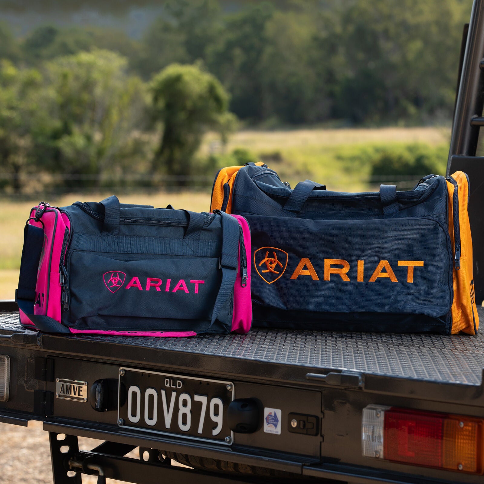 Two Ariat Gear Bag Combo (4-600/4-500) bags, one with pink accents and one with orange, rest on a vehicle’s back with a Queensland plate visible. Both feature padded shoulder straps for comfortable carrying. Brand: Ariat.