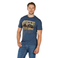 A man with short hair and a trimmed beard wears a Bullzye Mens Muster S/S Tee (B6W1503510) in Blue Steel, featuring bison, a Jeep, and the slogan "BUSTIN' THE HUB." He poses against a plain white background with one hand in his pocket.