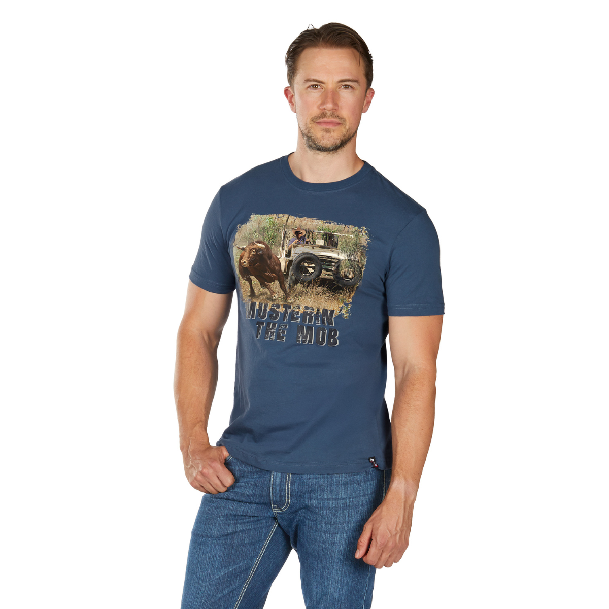 A man with short hair and a trimmed beard wears a Bullzye Mens Muster S/S Tee (B6W1503510) in Blue Steel, featuring bison, a Jeep, and the slogan "BUSTIN' THE HUB." He poses against a plain white background with one hand in his pocket.