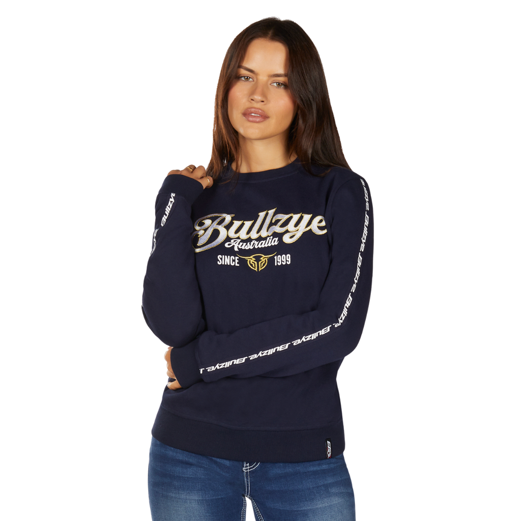 A woman with long brown hair wears a Bullzye Womens Ellie Crew Jumper (B6W2539532) in navy and blue jeans as she poses against a plain white background.