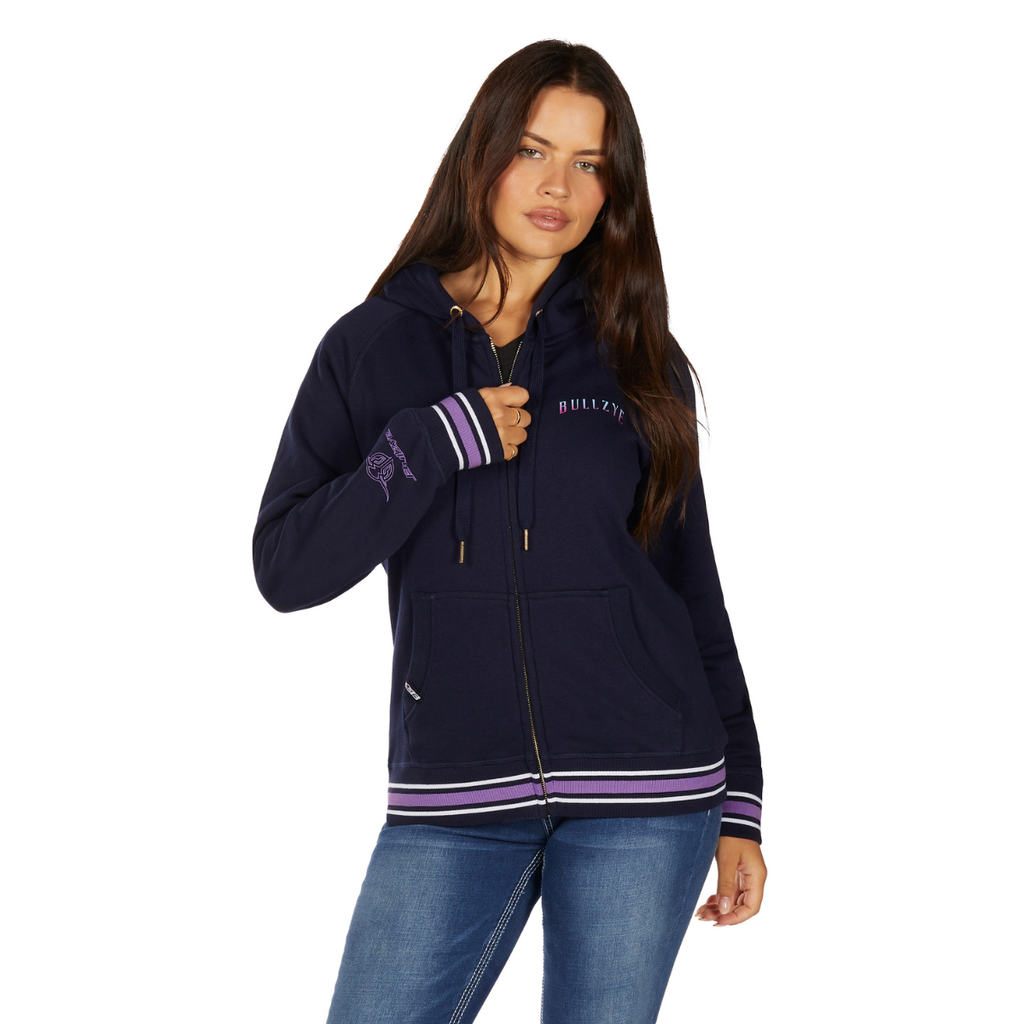 A woman with long brown hair wears the Bullzye Womens Jemma Zip Up Hoodie (B6W2505534) in navy, featuring striped cuffs and hem, paired with blue jeans. She faces forward, one hand on the hoodie strings and the other by her side.