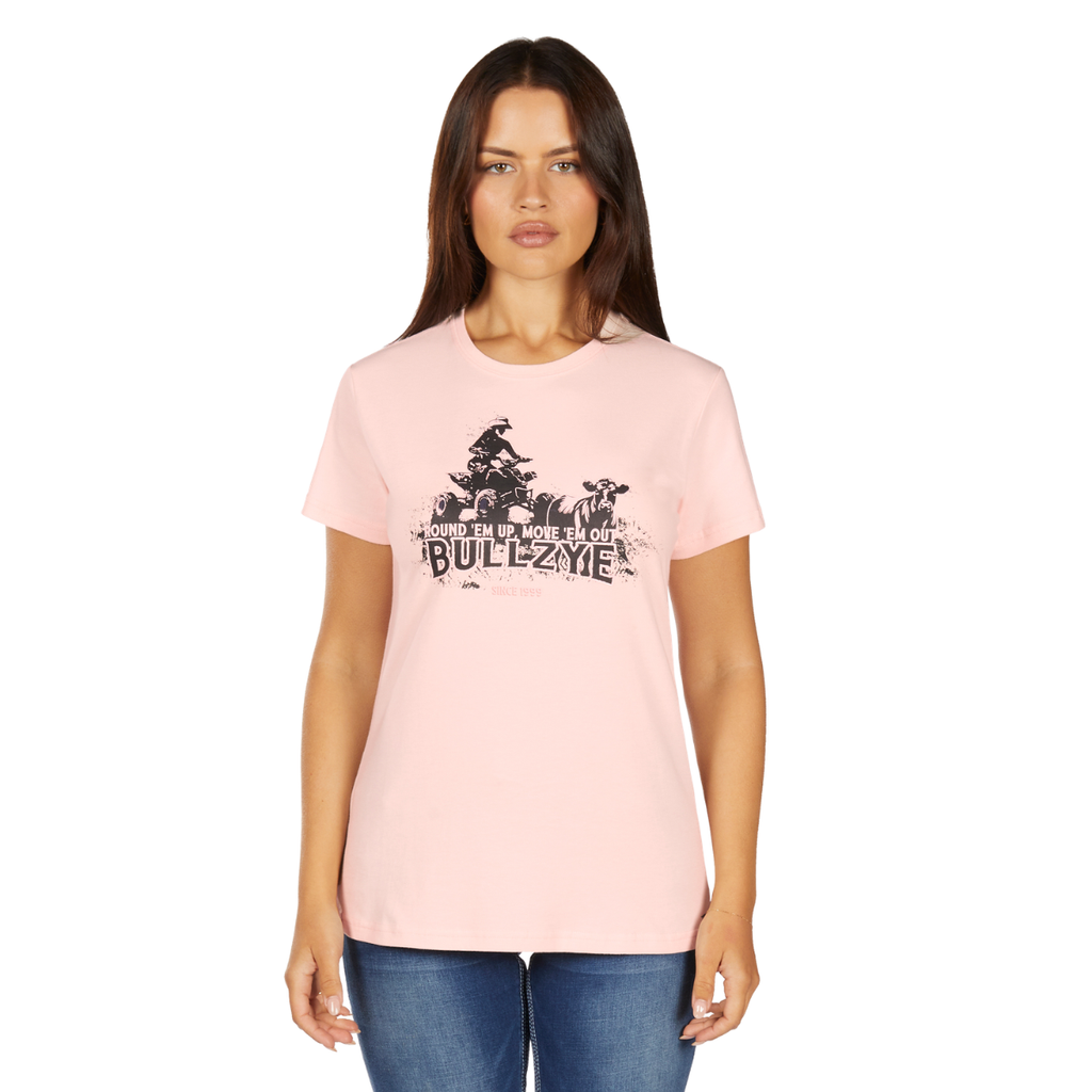 A woman with long brown hair wears the Bullzye Womens Lucy S/S Tee (B6W2503525) in blush, featuring a graphic and the phrase "Round 'em up, move 'em out, BULLZYE," while standing forward in blue jeans.
