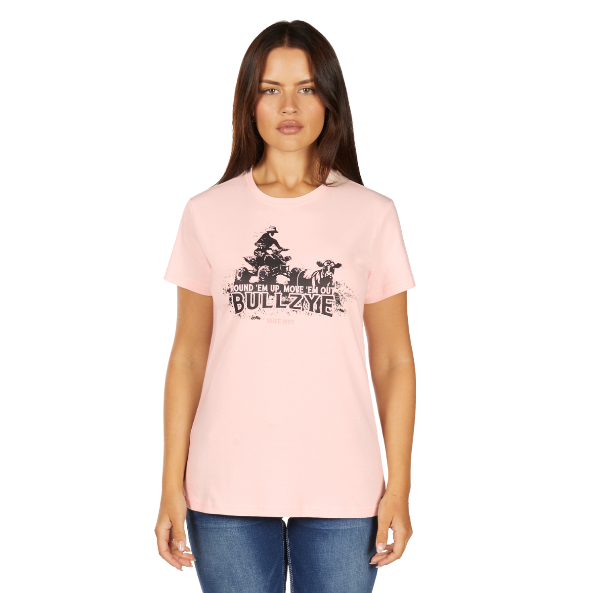 A woman with long brown hair wears the Bullzye Womens Lucy S/S Tee (B6W2503525) in blush, featuring a graphic and the phrase "Round 'em up, move 'em out, BULLZYE," while standing forward in blue jeans.