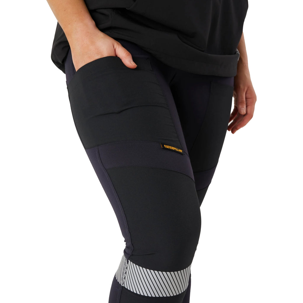 A close up of a woman from the waist down wearing navy blue CAT Women's Taped Work Stretch Leggings (1810096) with hi vis taping on the lower legs, showing the pocket detail, paired with a black tee on a white background.