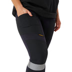 A close up of a woman from the waist down wearing navy blue CAT Women's Taped Work Stretch Leggings (1810096) with hi vis taping on the lower legs, showing the pocket detail, paired with a black tee on a white background.