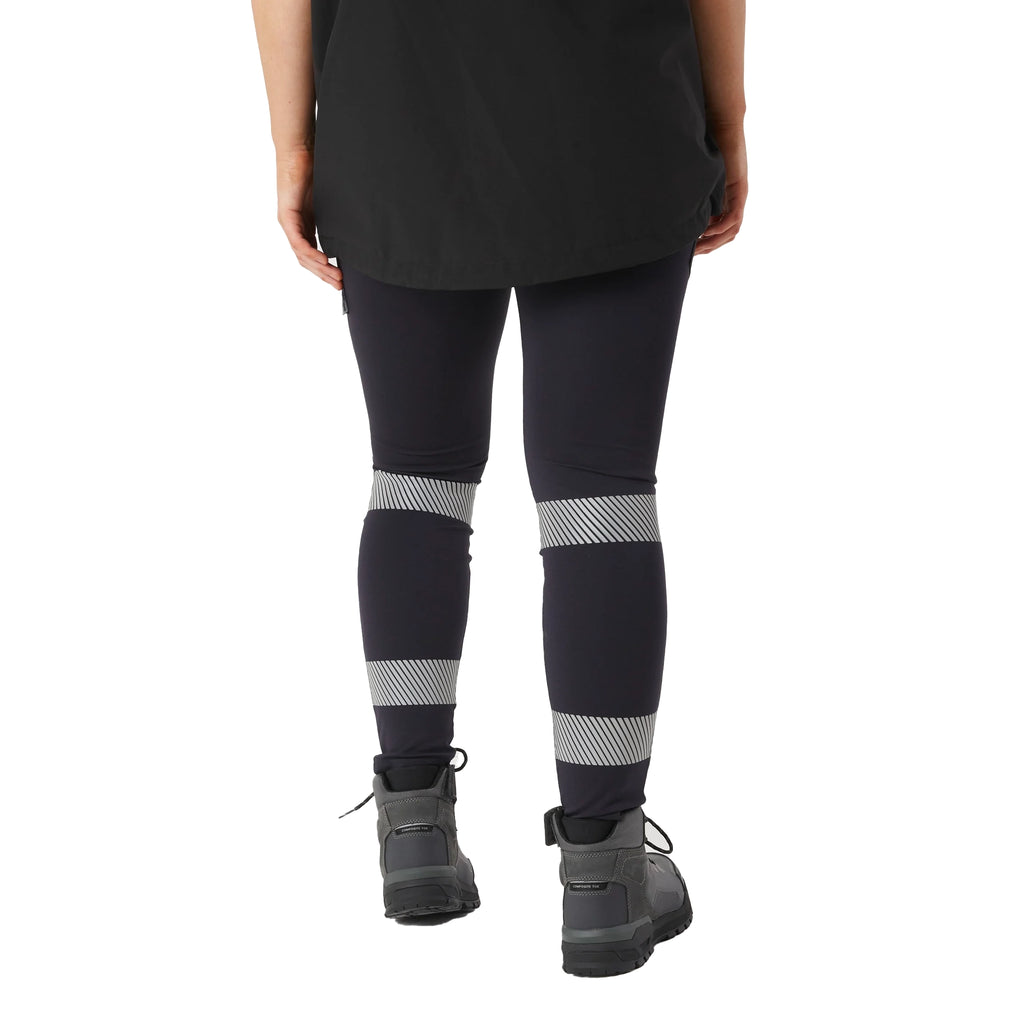 A woman viewed from behind, from the waist down wearing navy blue CAT Women's Taped Work Stretch Leggings (1810096) with hi vis taping on the lower legs, paired with a black tee and honey coloured work boots, on a white background.