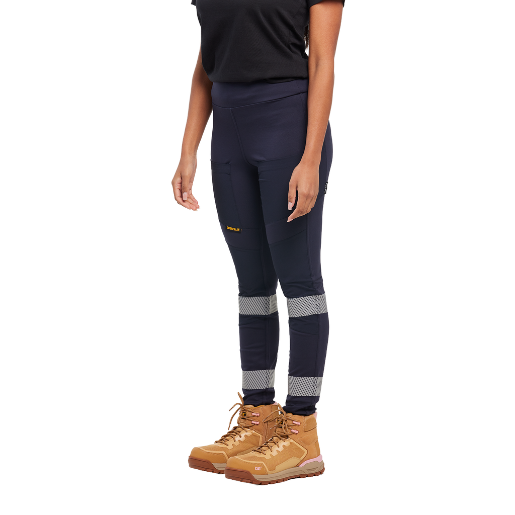 A woman from the waist down wearing navy blue CAT Women's Taped Work Stretch Leggings (1810096) with hi vis taping on the lower legs, paired with a black tee and honey coloured work boots, on a white background.