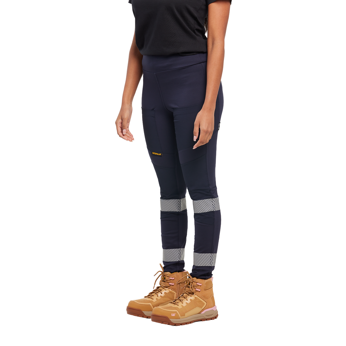 A woman from the waist down wearing navy blue CAT Women's Taped Work Stretch Leggings (1810096) with hi vis taping on the lower legs, paired with a black tee and honey coloured work boots, on a white background.