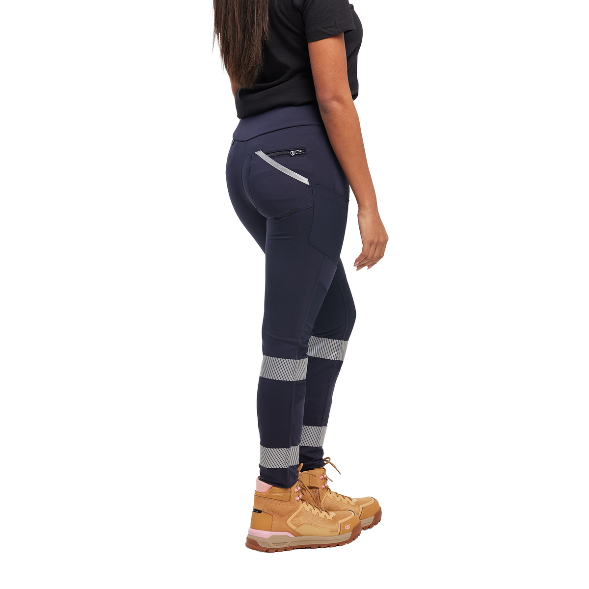 A woman viewed from 3-4 back from the waist down wearing navy blue CAT Women's Taped Work Stretch Leggings (1810096) with hi vis taping on the lower legs, paired with a black tee and honey coloured work boots, on a white background.