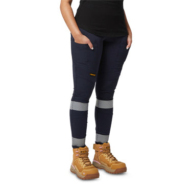 A woman from the waist down wearing navy blue CAT Women's Taped Work Stretch Leggings (1810096) with hi vis taping on the lower legs, showing the pocket detail, paired with a black tee and honey coloured work boots on a white background.