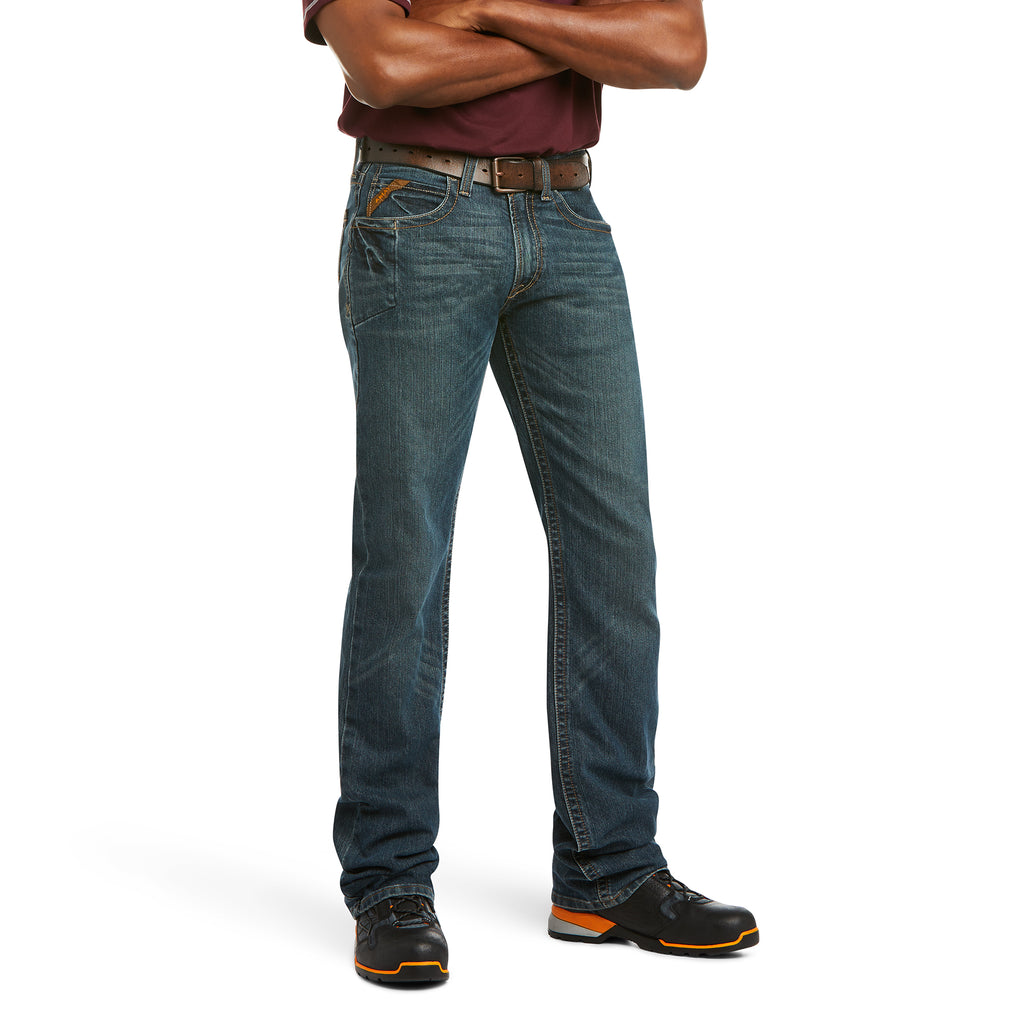 A person with crossed arms wears a maroon shirt, Ariat Rebar M5 Jeans (10016222) Ironside by Ariat, a brown belt, and black shoes with orange accents. The face is not visible in the image.