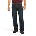 A man, face not visible, stands against a white background wearing a maroon shirt, brown belt, black work boots, and Ariat Rebar M5 Jeans (10016223) Blackstone by Ariat.