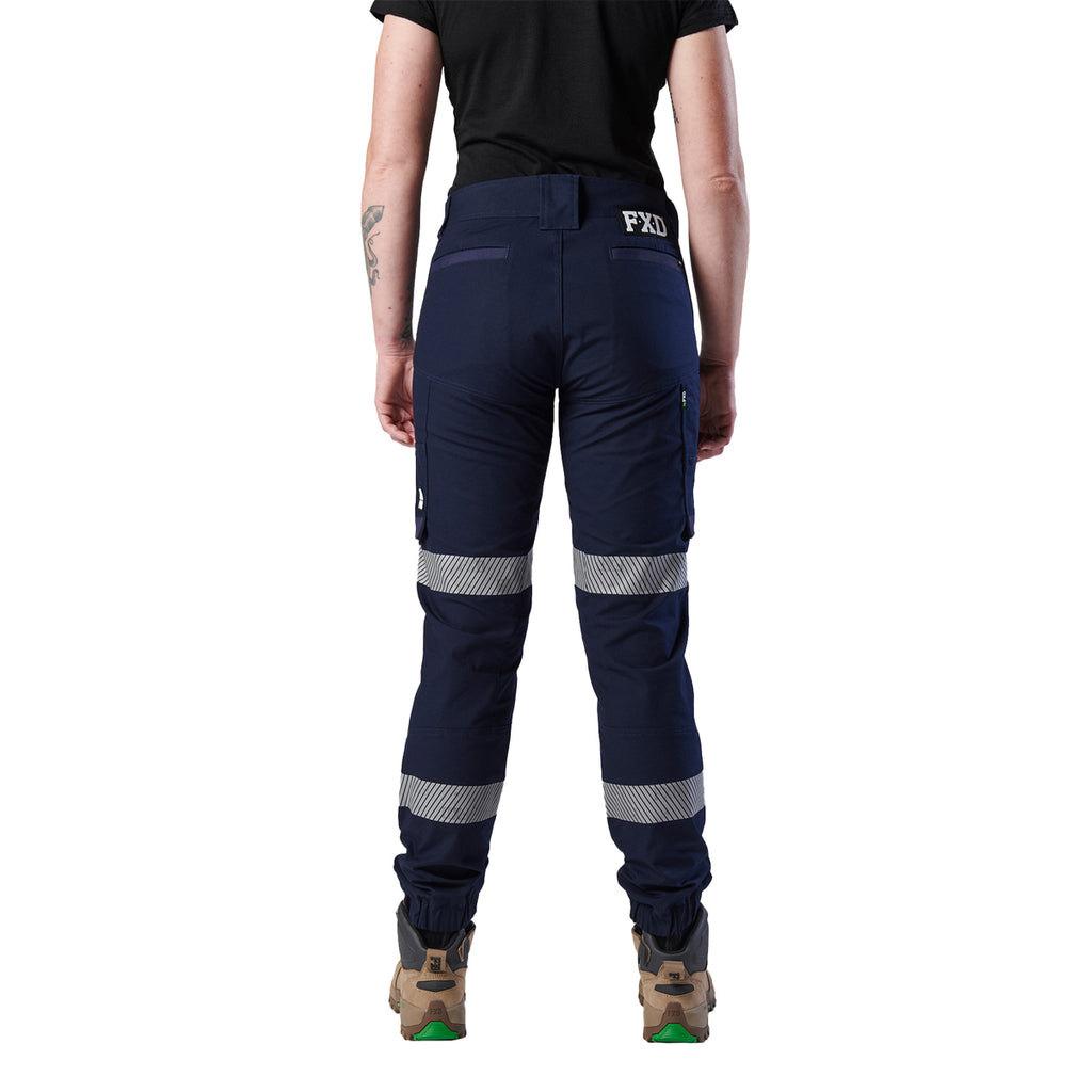 A rear view of a woman wearing a pair of navy blue FXD Women's Cuffed Taped Work Pants (WP-8WT) with reflective silver-grey bands on the legs, paired with a black t-shirt and stone coloured boots, against a plain white background.
