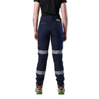 A rear view of a woman wearing a pair of navy blue FXD Women's Cuffed Taped Work Pants (WP-8WT) with reflective silver-grey bands on the legs, paired with a black t-shirt and stone coloured boots, against a plain white background.