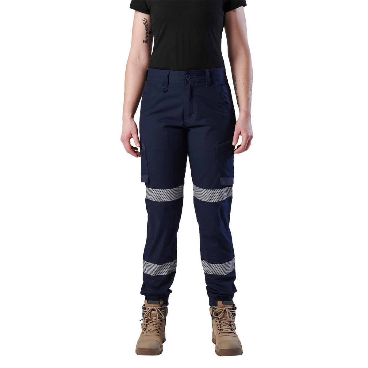 A woman wearing a pair of navy blue FXD Women's Cuffed Taped Work Pants (WP-8WT) with reflective silver-grey bands on the legs, paired with a black t-shirt and stone coloured boots, against a plain white background.