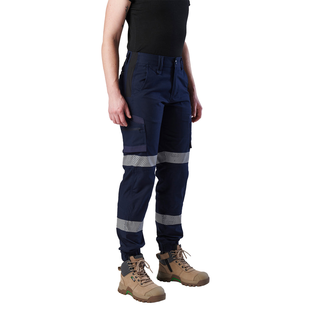 A 3-4 front view of a woman wearing a pair of navy blue FXD Women's Cuffed Taped Work Pants (WP-8WT) with reflective silver-grey bands on the legs, paired with a black t-shirt and stone coloured boots, against a plain white background.