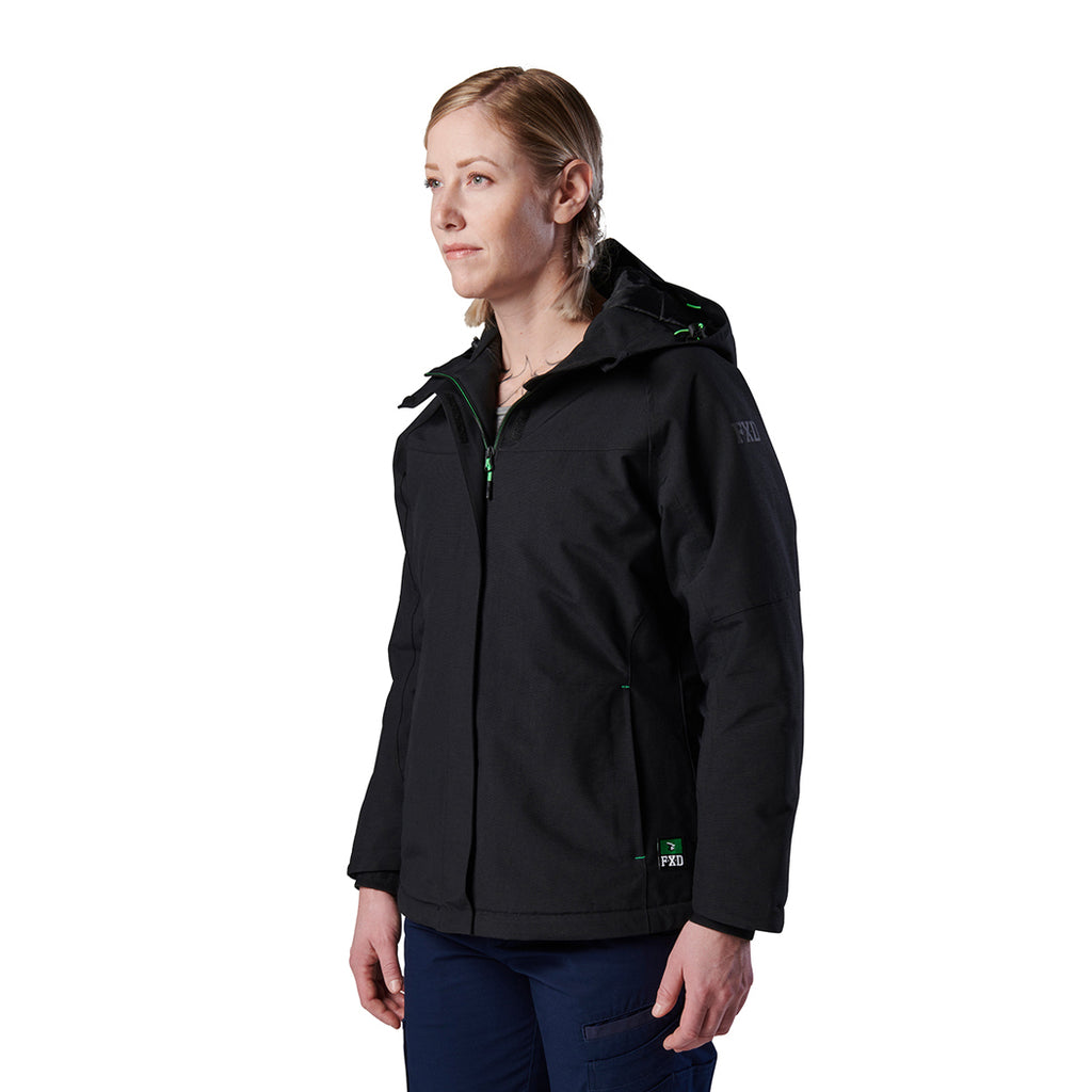A woman modelling a black hooded FXD Women's WO-1W Insulated Waterproof Jacket (FX12407001) with green zipper accents and a small green logo, paired with navy pants, photographed against a plain white studio background.