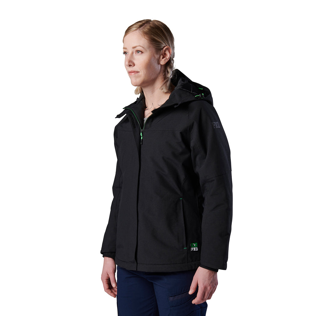A woman modelling a black hooded FXD Women's WO-1W Insulated Waterproof Jacket (FX12407001) with green zipper accents and a small green logo, paired with navy pants, photographed against a plain white studio background.