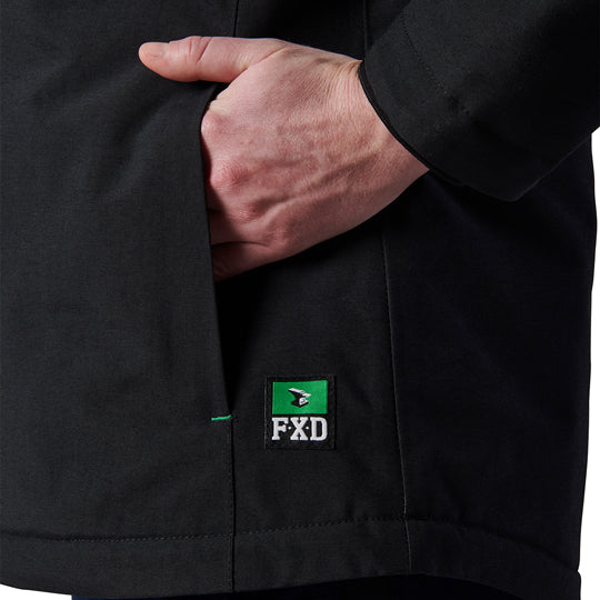 Close-up of a woman modelling a black hooded FXD Women's WO-1W Insulated Waterproof Jacket (FX12407001), with a hand tucked into a side pocket and a small green and white FXD logo patch near the hem.