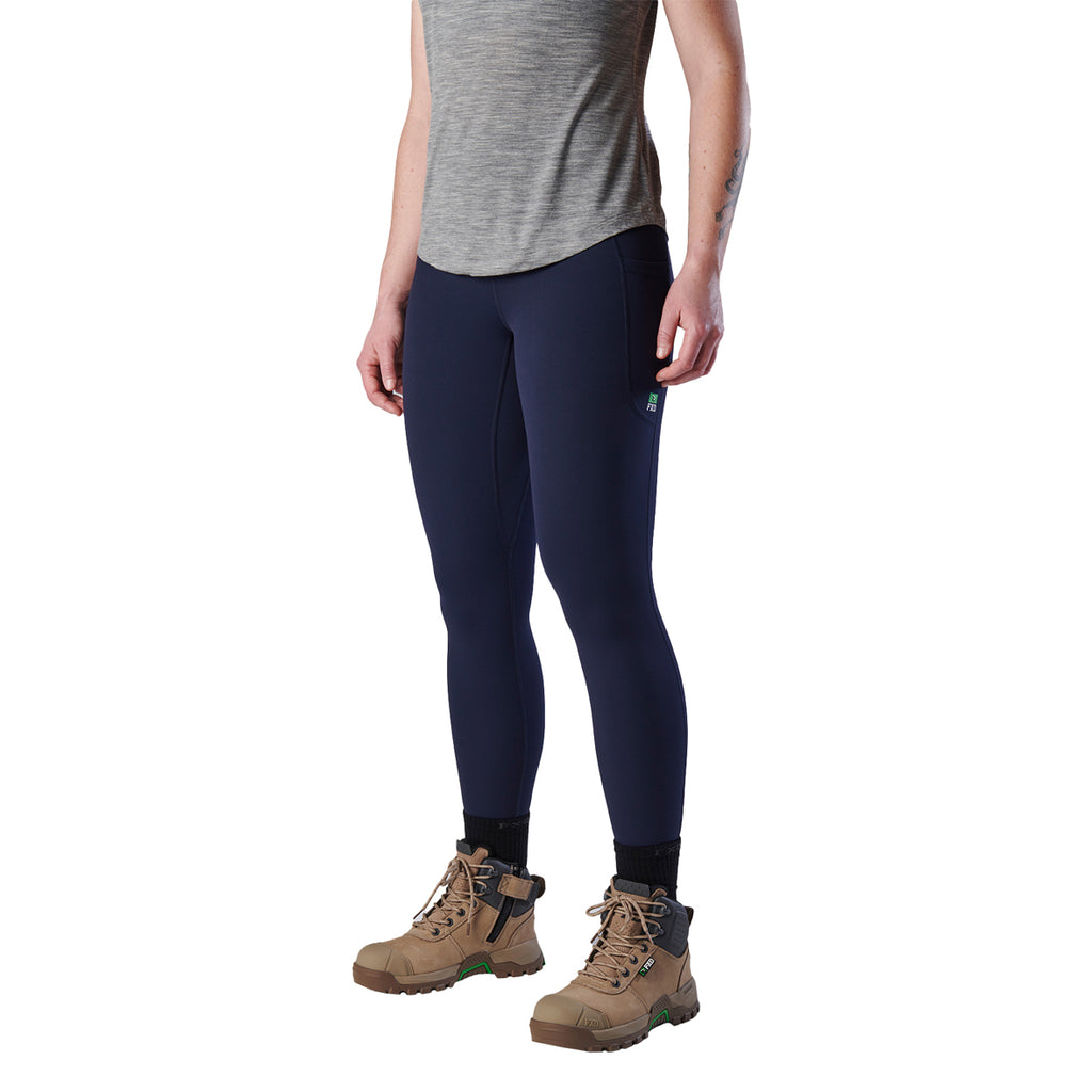 A woman viewed from behind modelling a pair of navy blue FXD Women's Work Leggings (WP-9W) paired with a grey tee and stone coloured work boots on a white background.
