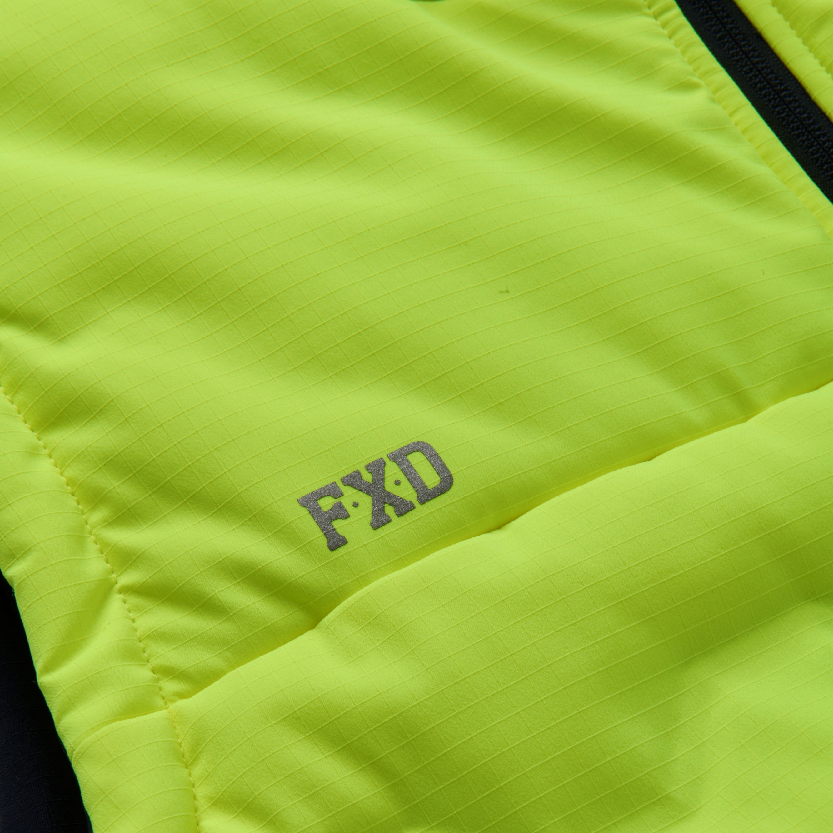 The FXD Mens WO-6 Puffer Vest (FX02407005) in yellow is a sleeveless, water-repellent insulated vest with black side panels, black zipper, high collar, padded warmth, and FXD branding on the chest and lower left hem.