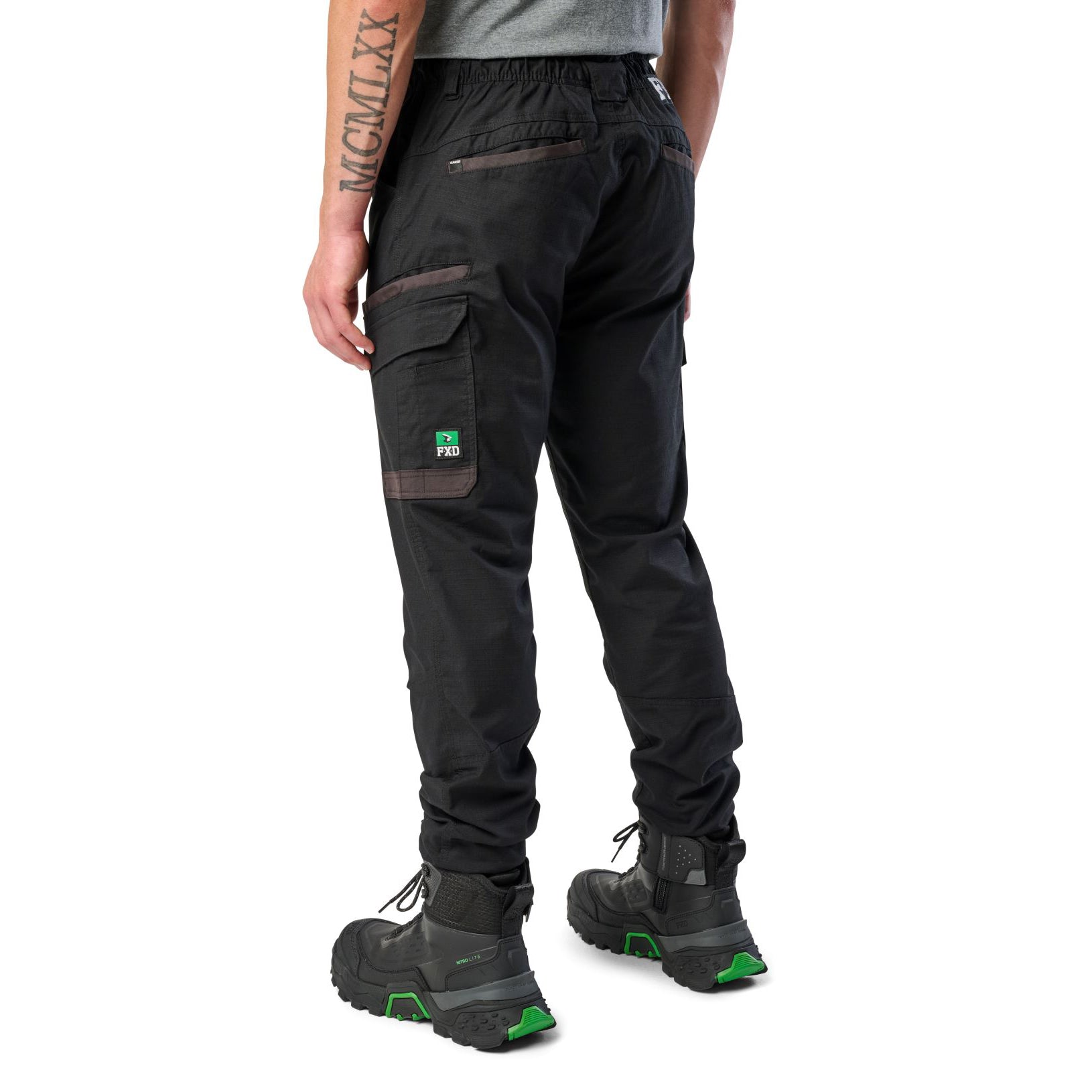 A man wearing black cargo style FXD WP-12C Elastic Waist Stretch Ripstop Cuffed Work Pants (FX02506027) with a drawstring waist and black work boots, shown from the waist down on a plain white studio background, 3-4 back view.