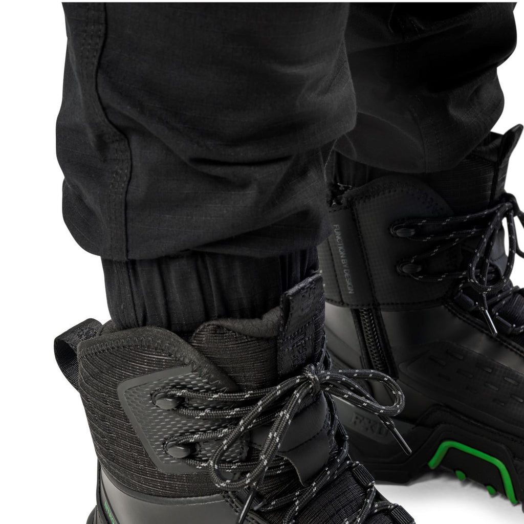 Cuff detail of a pair of black cargo style FXD WP-12C Elastic Waist Stretch Ripstop Cuffed Work Pants (FX02506027) and black work boots, on a plain white studio background.