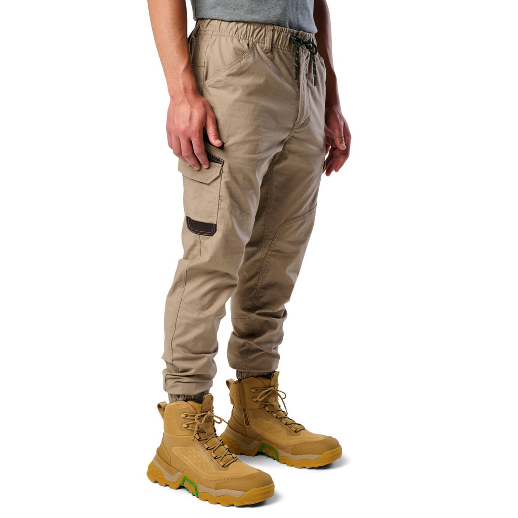 A 3-4 front view of a man wearing khaki cargo style FXD WP-12C Elastic Waist Stretch Ripstop Cuffed Work Pants (FX02506027) with a drawstring waist and wheat work boots, shown from the waist down on a plain white studio background.