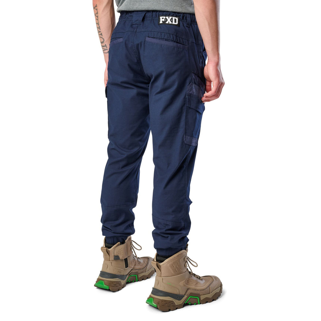 A 3-4 back view of a man wearing navy cargo style FXD WP-12C Elastic Waist Stretch Ripstop Cuffed Work Pants (FX02506027) with a drawstring waist and stone work boots, shown from the waist down on a plain white studio background.