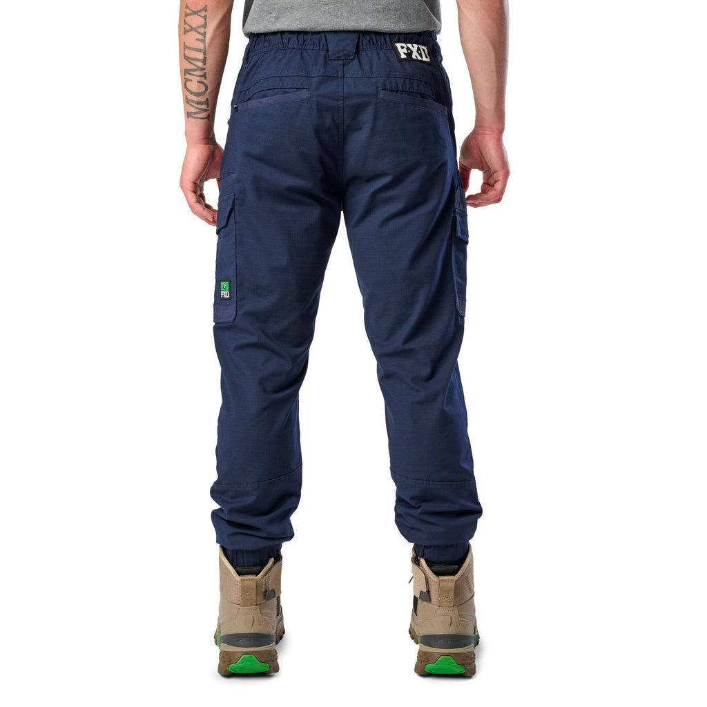 A back view of a man wearing navy cargo style FXD WP-12C Elastic Waist Stretch Ripstop Cuffed Work Pants (FX02506027) with a drawstring waist and stone work boots, shown from the waist down on a plain white studio background.