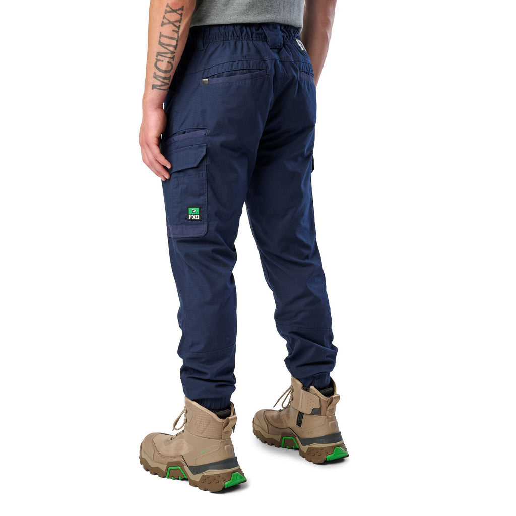 A 3-4 back view of a man wearing navy cargo style FXD WP-12C Elastic Waist Stretch Ripstop Cuffed Work Pants (FX02506027) with a drawstring waist and stone work boots, shown from the waist down on a plain white studio background.