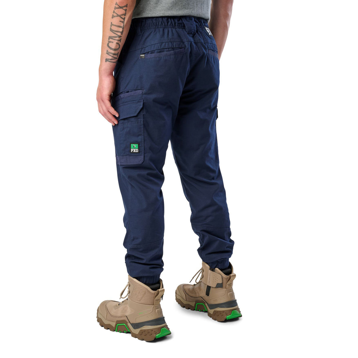 A 3-4 back view of a man wearing navy cargo style FXD WP-12C Elastic Waist Stretch Ripstop Cuffed Work Pants (FX02506027) with a drawstring waist and stone work boots, shown from the waist down on a plain white studio background.