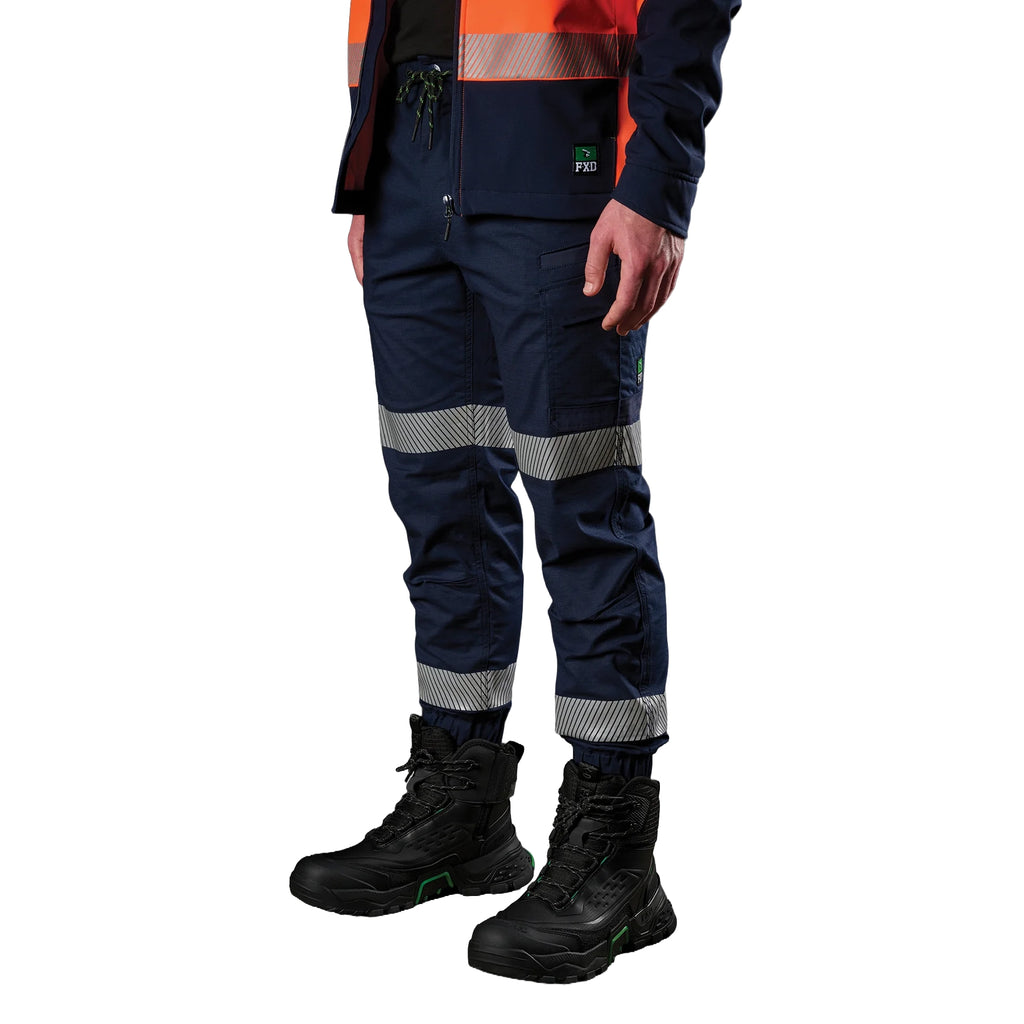 A 3-4 view of a man wearing navy FXD Men's WP-12CT Elastic Waist Stretch Ripstop Cuffed Taped Work Pants (FX02606029) with reflective tape at the knees and cuffs, paired with a matching navy and orange high-vis jacket and black boots, on a white background.