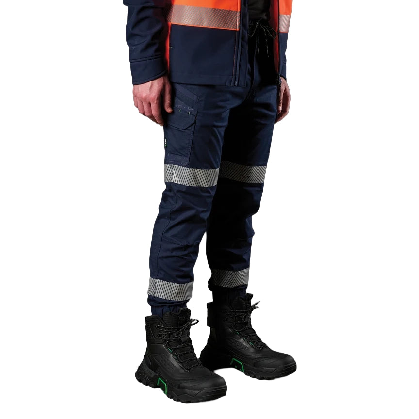 A 3-4 view of a man wearing navy FXD Men's WP-12CT Elastic Waist Stretch Ripstop Cuffed Taped Work Pants (FX02606029) with reflective tape at the knees and cuffs, paired with a matching navy and orange high-vis jacket and black boots, on a white background.