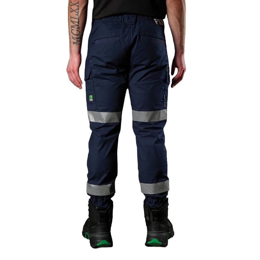A man shown from behind wearing navy FXD Men's WP-12CT Elastic Waist Stretch Ripstop Cuffed Taped Work Pants (FX02606029) with reflective tape at the knees and cuffs, paired with a matching navy and orange high-vis jacket and black boots, on a white background.