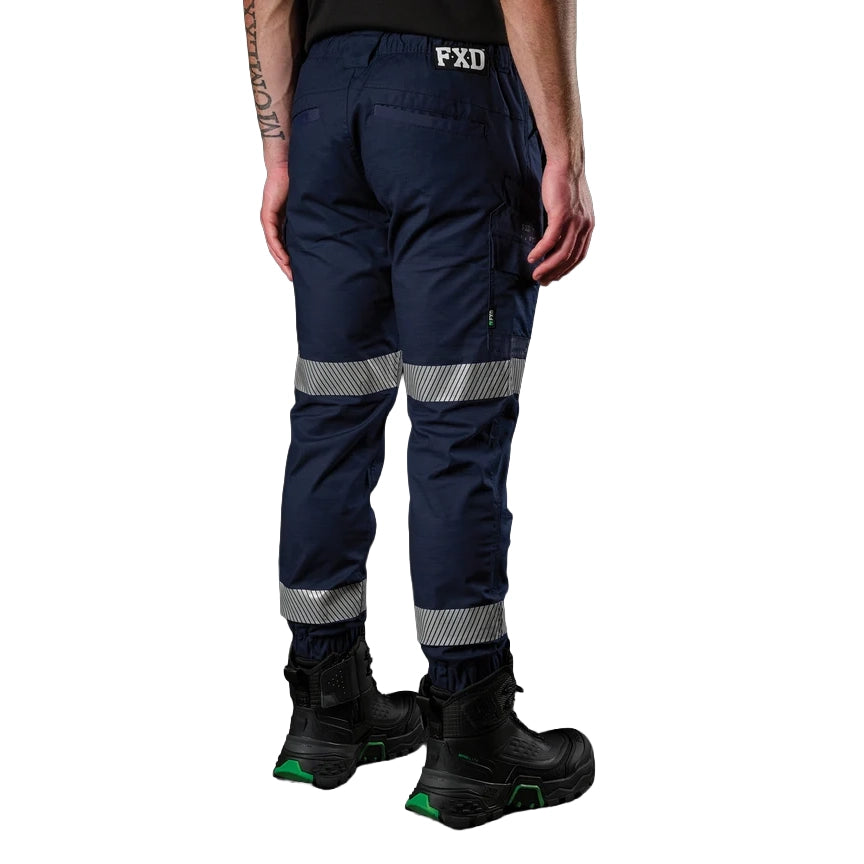 A 3-4 view of a man wearing navy FXD Men's WP-12CT Elastic Waist Stretch Ripstop Cuffed Taped Work Pants (FX02606029) with reflective tape at the knees and cuffs, paired with a matching navy and orange high-vis jacket and black boots, on a white background.