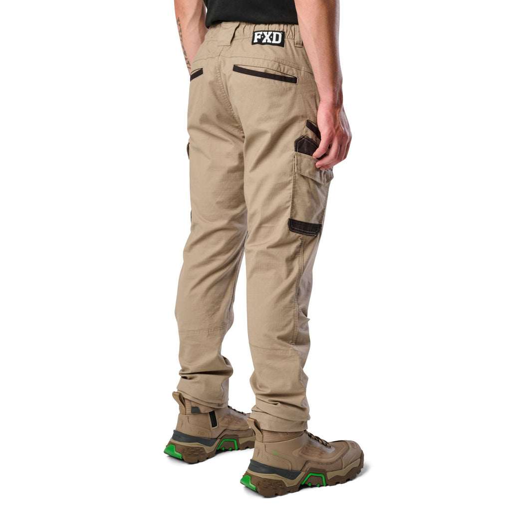 A 3-4 back view of a man wearing khaki cargo style FXD Men's WP-12 Elastic Waist Stretch Ripstop Work Pants (FX02506026) with stone coloured work boots on a white background.